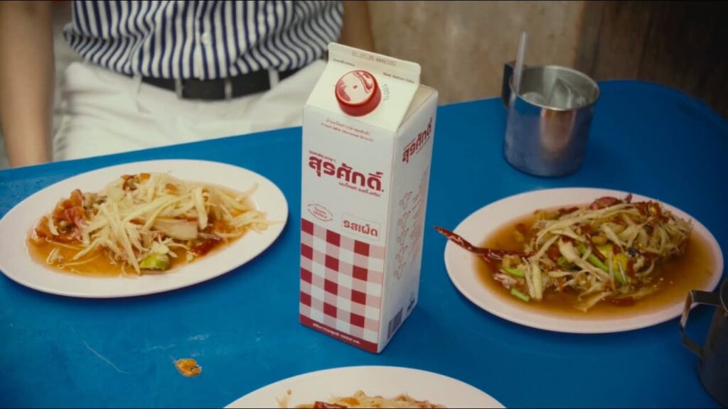 กล่องนมตราสุรศักดิ์ รสเผ็ด ลายตารางขาวแดง วางอยู่กลางโต๊ะอาหารสีน้ำเงิน ล้อมรอบด้วยจานส้มตำและแก้วน้ำแข็ง