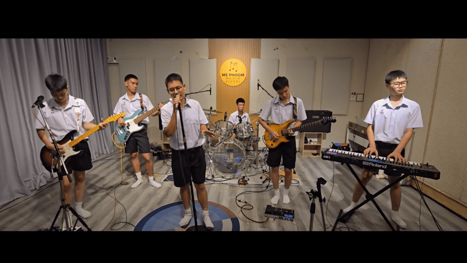 วงดนตรีนักเรียนชาย 6 คนในชุดเครื่องแบบ กำลังแสดงดนตรีและร้องเพลงคัฟเวอร์ในห้องซ้อมที่ได้มาตรฐานของโรงเรียนดนตรี Mephoom School of Music ประกอบด้วยนักร้องนำ มือกีตาร์ มือเบส มือกลอง และมือคีย์บอร์ด